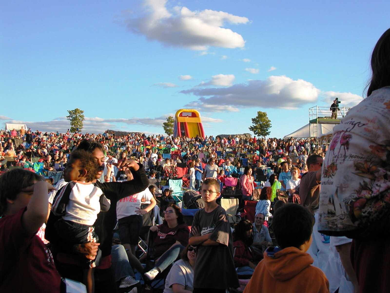 Large crowd seated on a hill at an outdoor festival with a colorful inflatable slide in the distance under a blue sky.