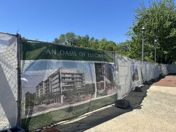 Construction site barrier with a banner reading 'AN OASIS OF ELEGANCE' showing a rendering of a modern building, trees and blue sky in the background.