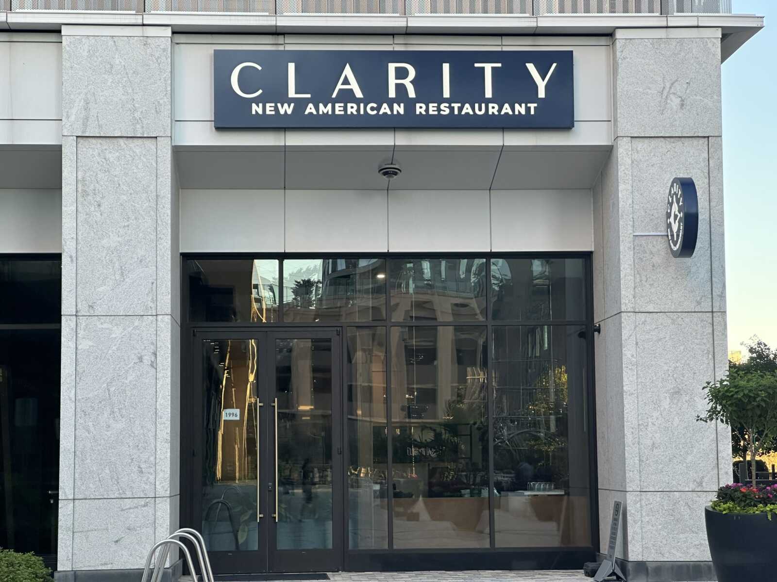 Facade of a modern restaurant entrance with a dark sign reading CLARITY NEW AMERICAN RESTAURANT above glass doors.