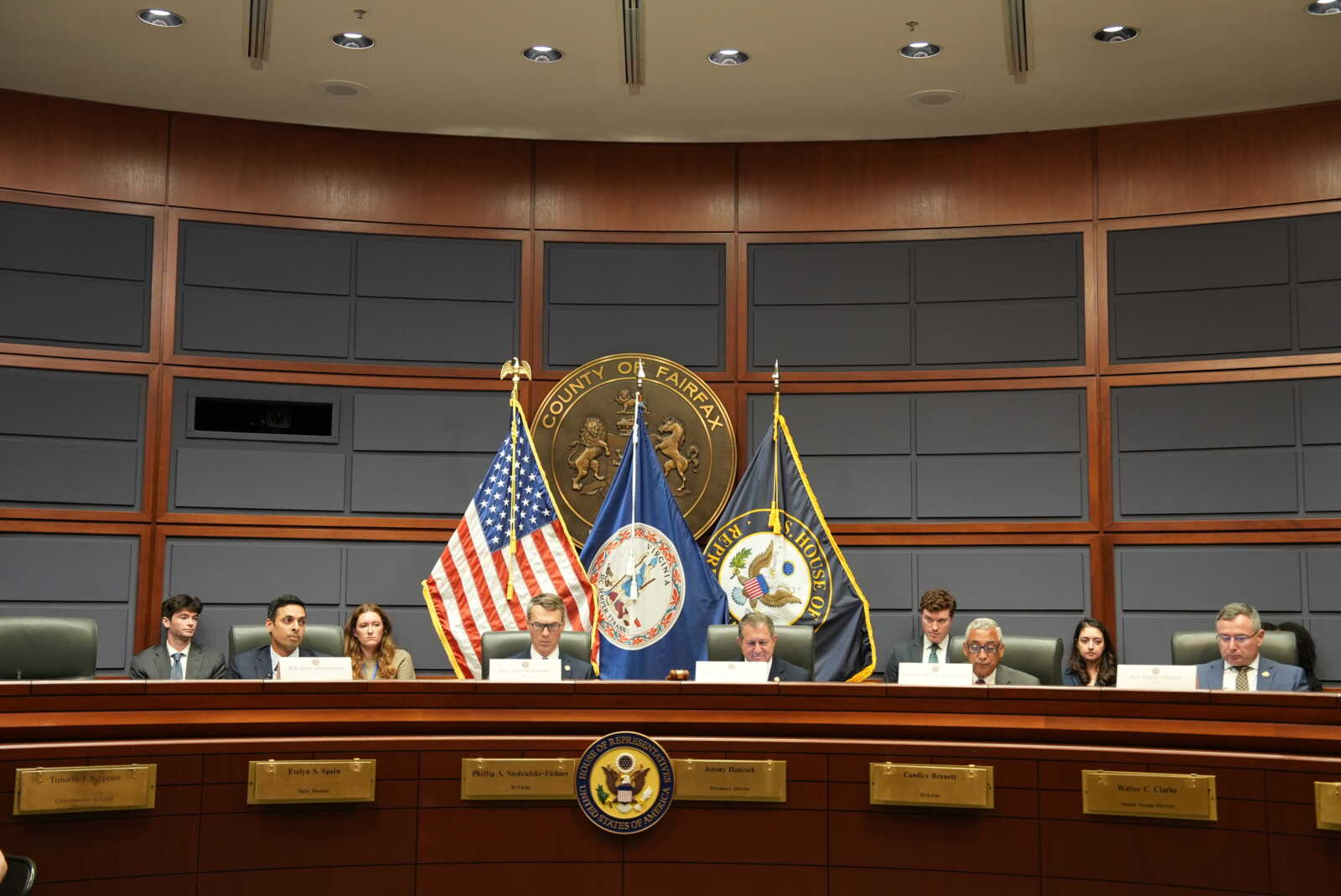 Panel of officials seated at a long dais in a wood-paneled chamber, with U.S. and state flags and a circular seal behind them, suggesting a formal government meeting.