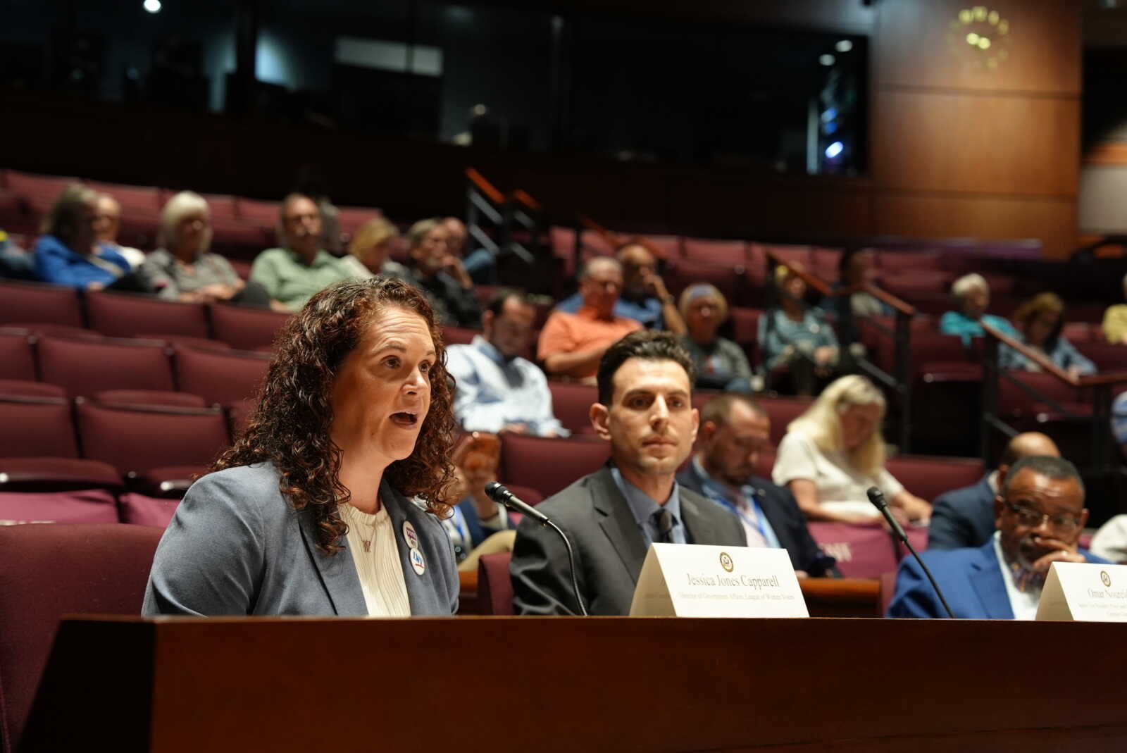 Woman with curly hair speaks into a microphone at a conference panel, seated at a table with name placards and an audience behind her.