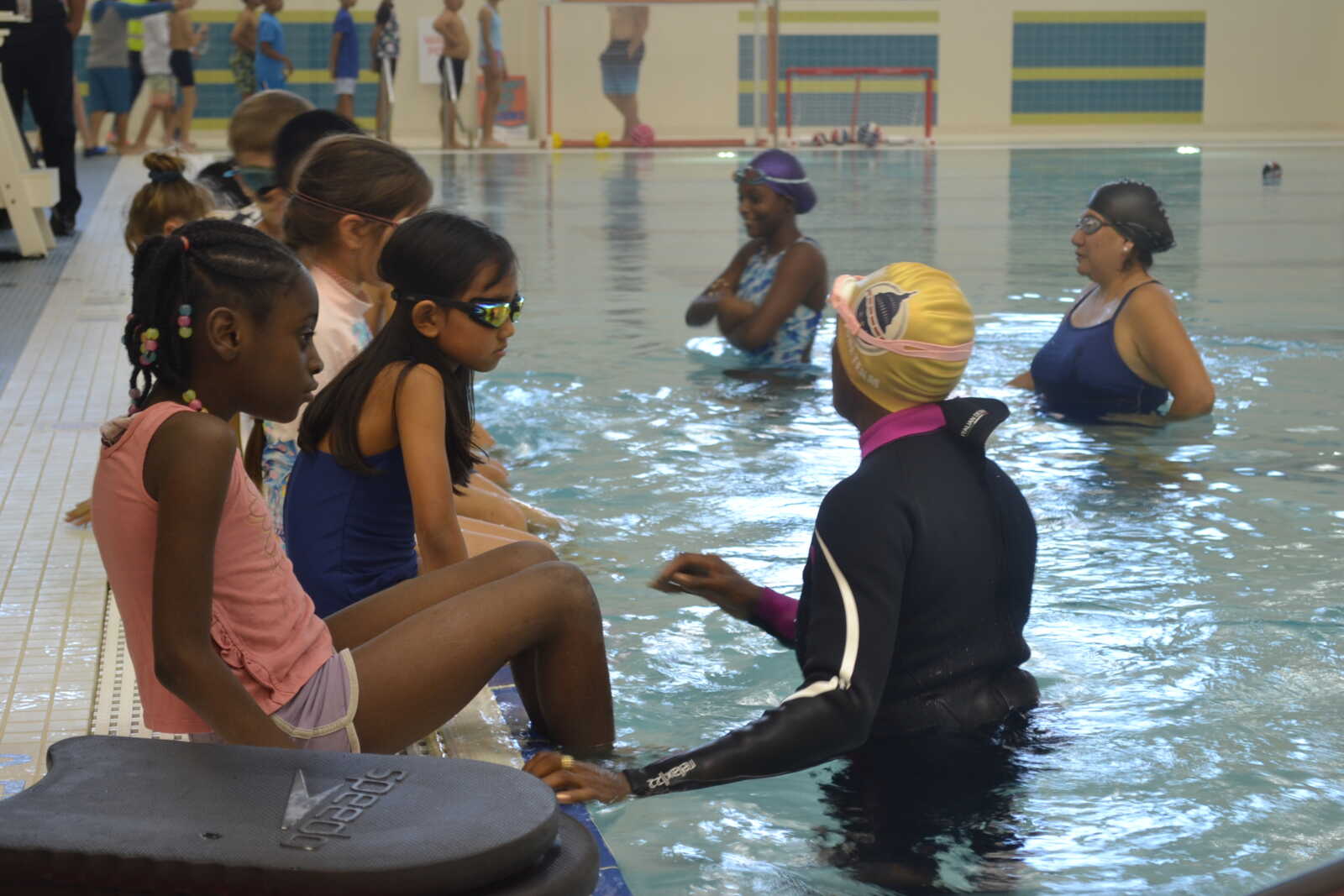 Group of children sitting at the pool edge watching an instructor in a wetsuit demonstrate in the water.