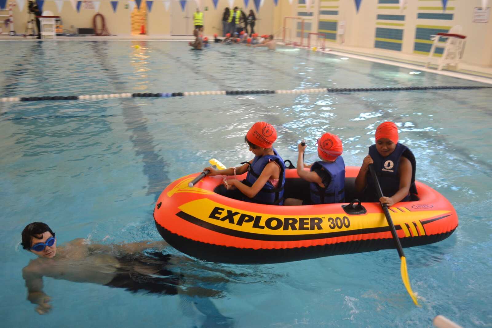 Three children wearing life jackets and orange swim caps sit in an orange Explorer 300 inflatable boat in a pool, paddling with oars while a swimmer nearby watches from the water.