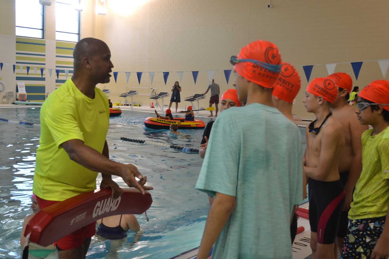 Lifeguard in neon shirt speaks to a group of boys wearing orange swim caps at pool edge during a class or safety drill, with a red rescue buoy in hand.