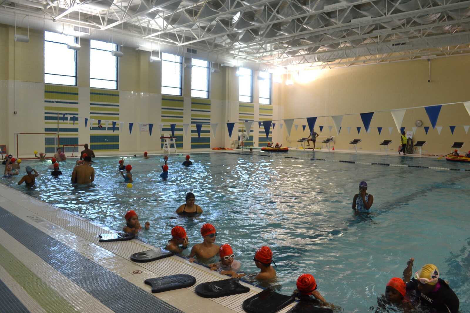 Indoor swimming pool scene with kids wearing red swim caps in the water, some adults nearby, and lane ropes with blue and white bunting overhead.