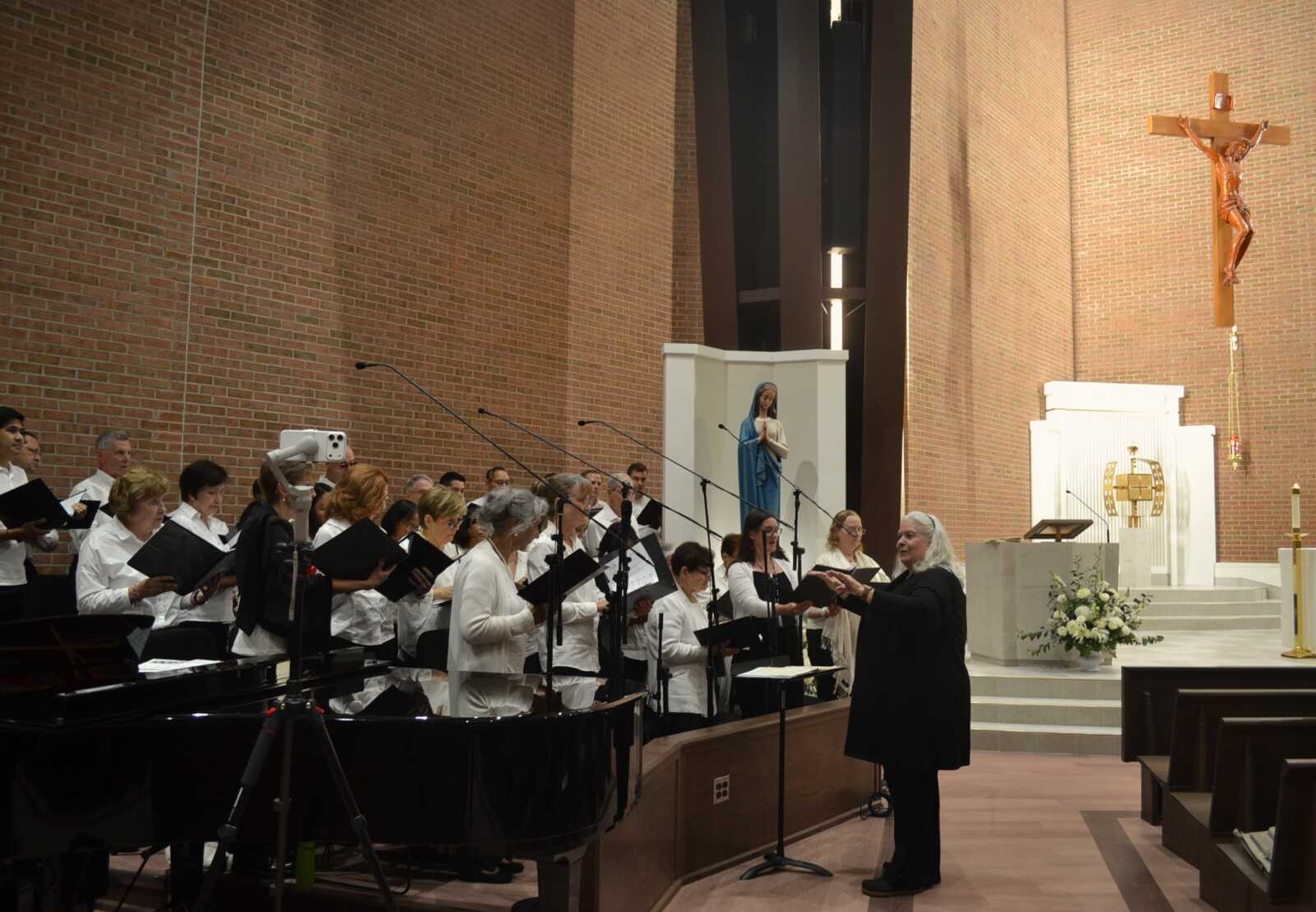 A choir of people in white shirts singing in a church, with a conductor guiding them near a piano and brick walls.