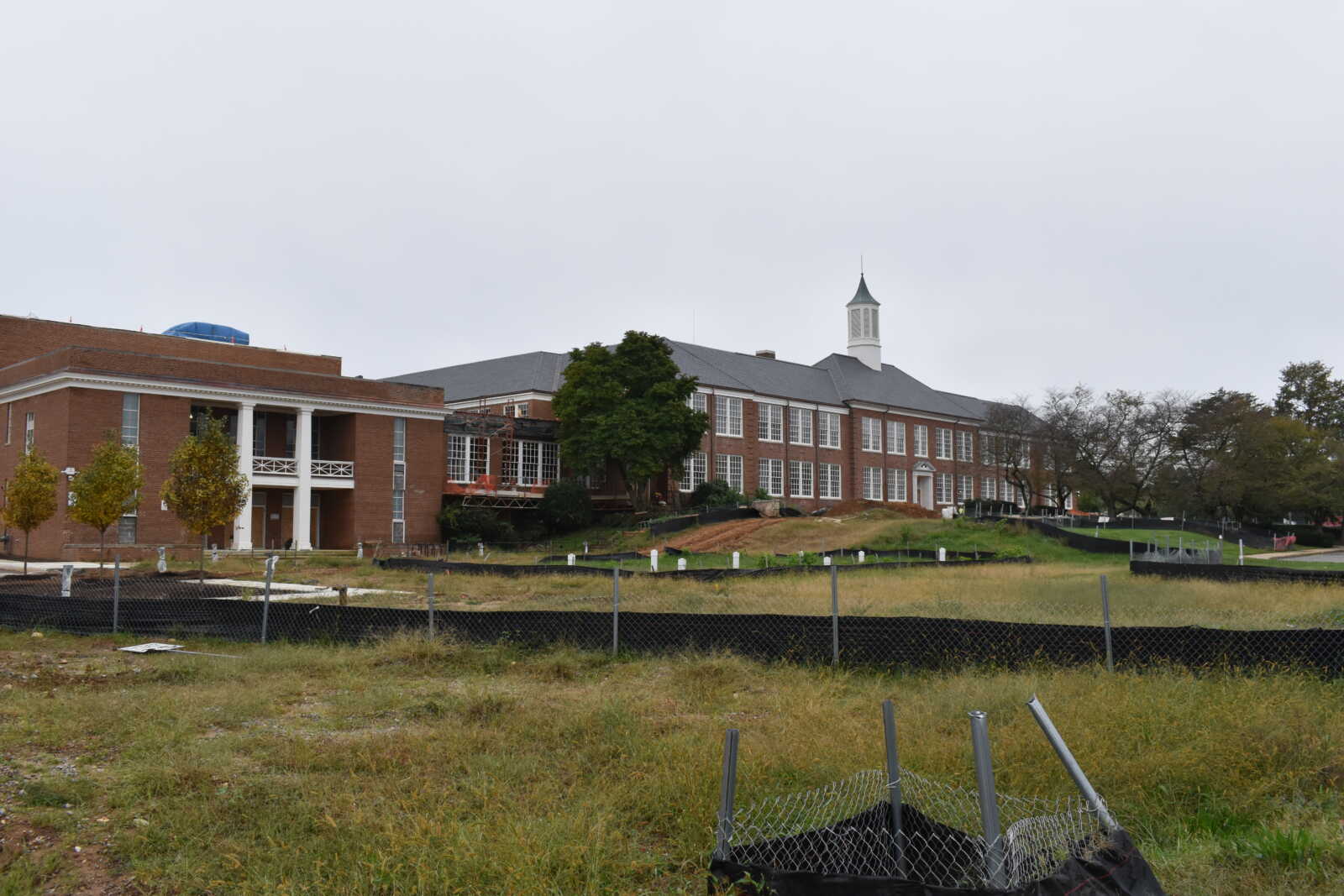 Brick school building with a white cupola on its roof, under construction, surrounded by a chain-link fence and a grassy field in the foreground