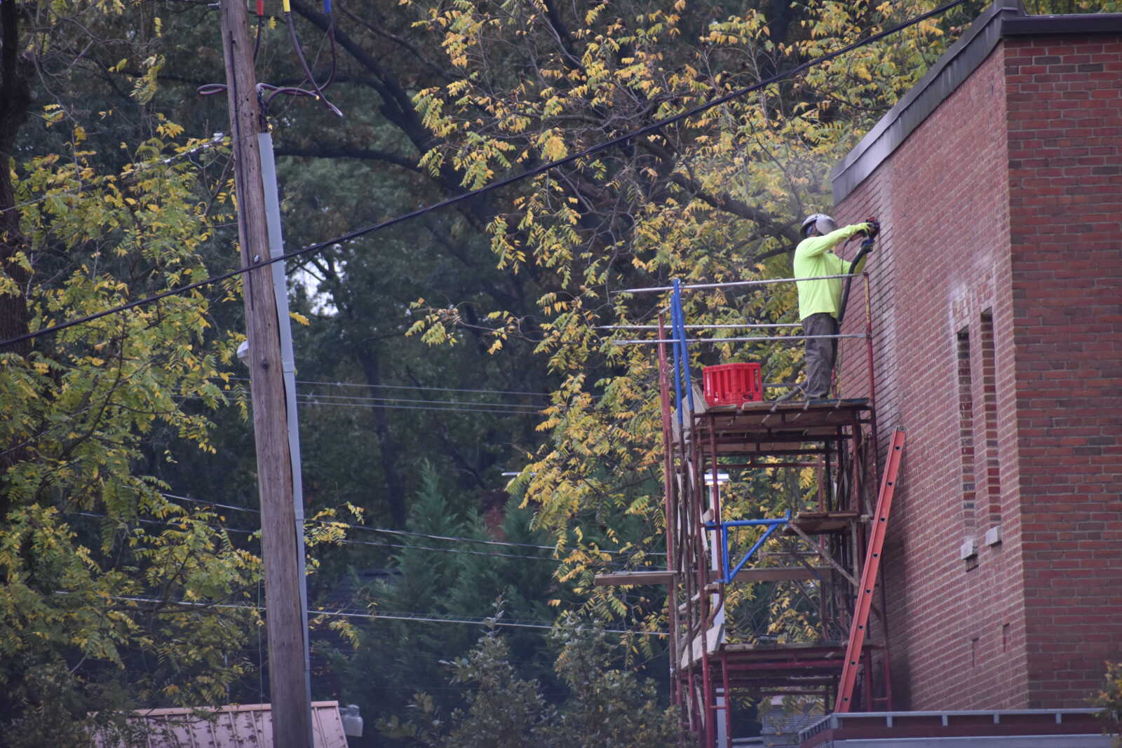 Construction worker in a bright lime shirt and hard hat on scaffolding beside a brick building, adjusting a tool.