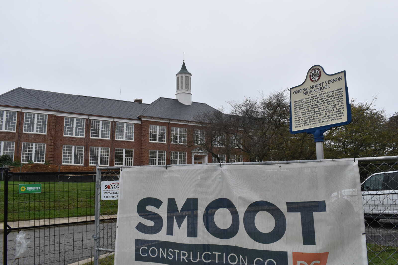 Construction site in front of a brick school building with a large SMOOT Construction banner on a chain-link fence in the foreground.