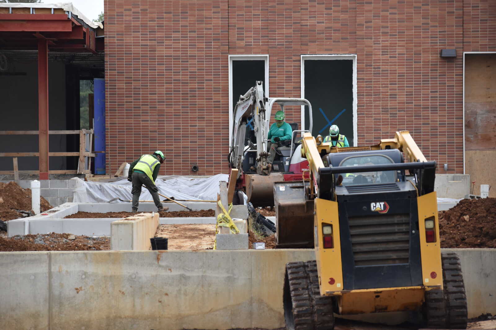 Construction site with workers in green safety vests and helmets operating a backhoe in front of a brick building.
