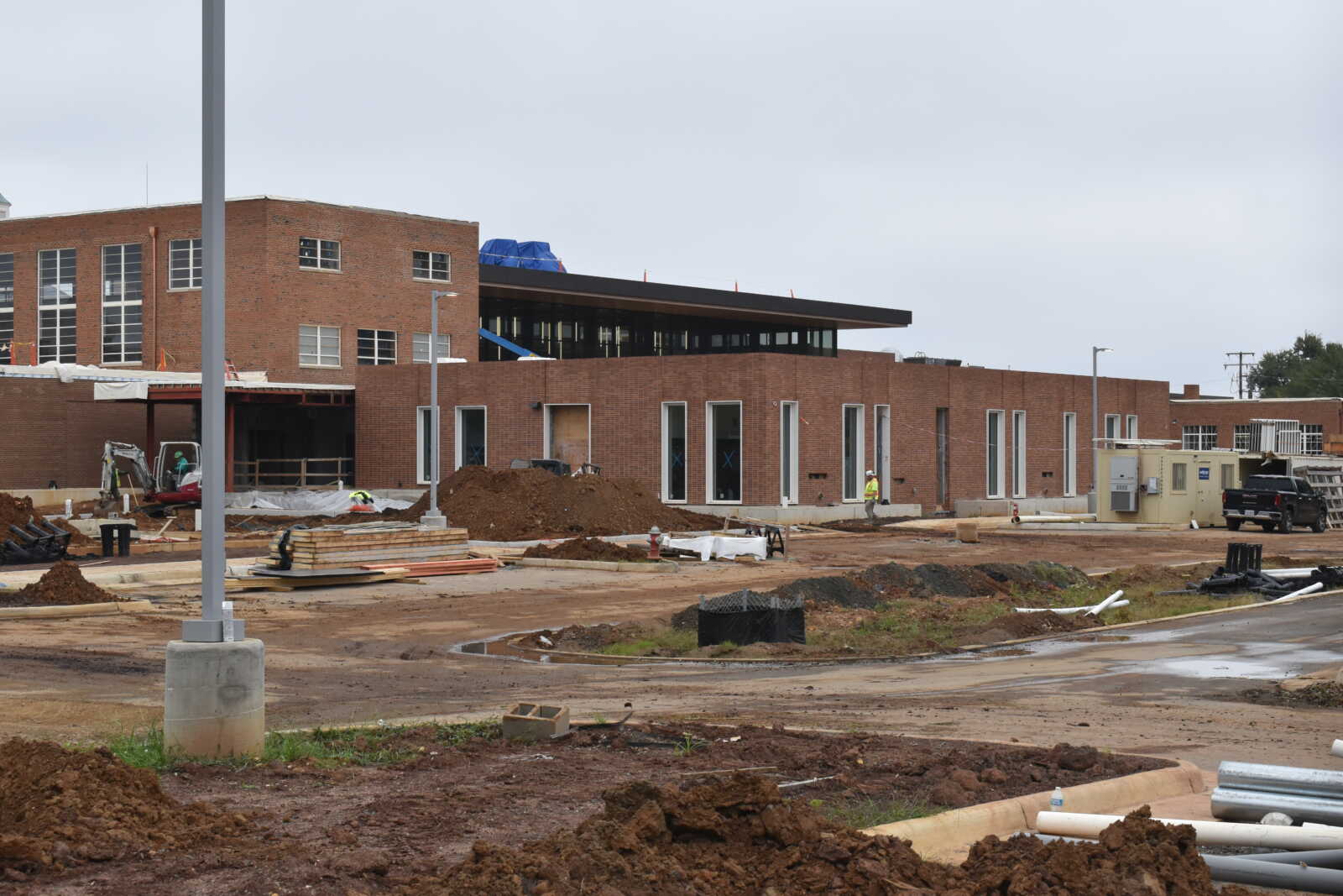 Construction site with a brick building under construction and dirt piles in the foreground; construction equipment and a black pickup truck visible to the right.