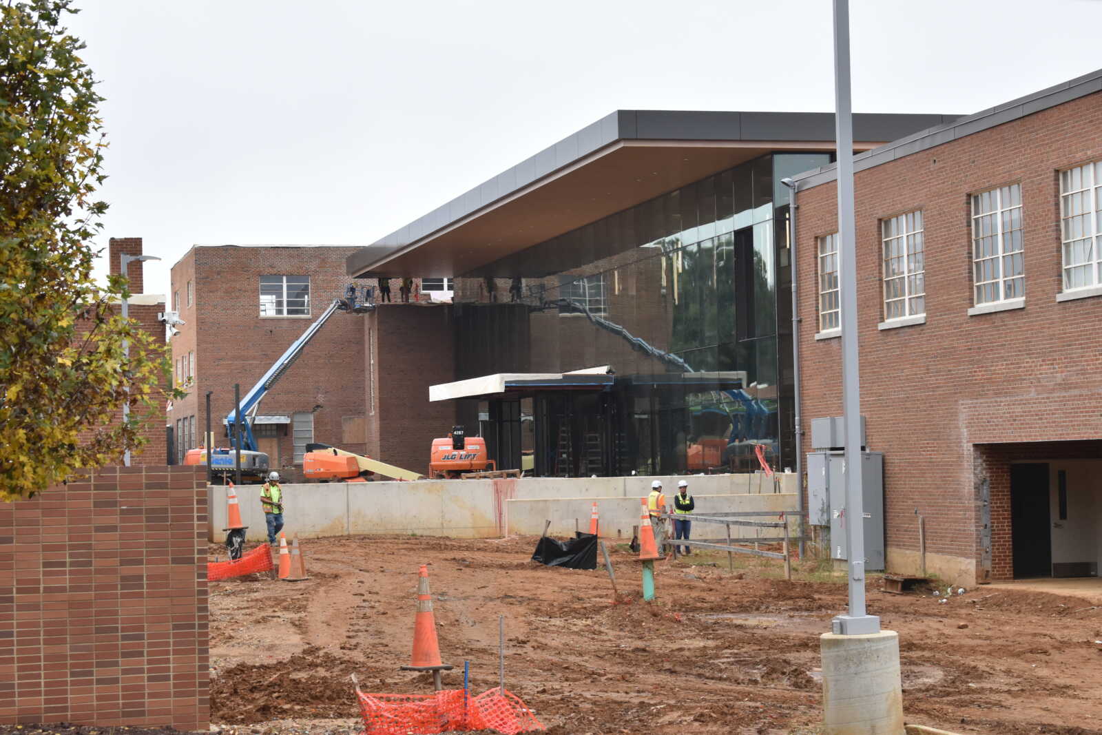 Construction site in front of a brick and glass building; workers with hard hats and orange safety barriers on a dirt lot.