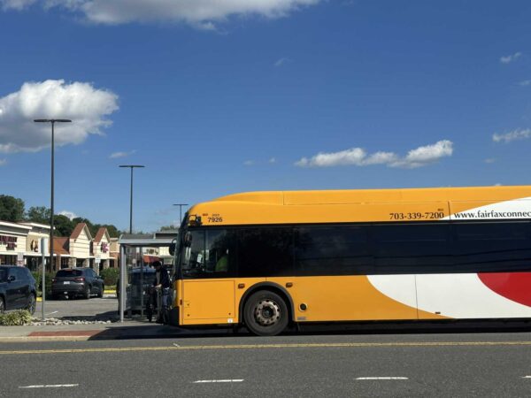 Yellow transit bus at a curbside stop in a shopping center parking lot, with storefronts and parked cars in the background under a blue sky.
