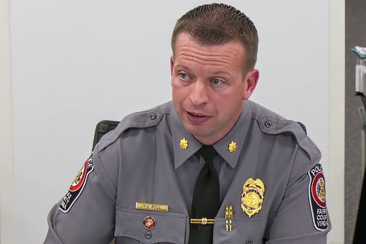 Police officer in a gray uniform with gold badge and nameplate, seated and speaking in a meeting room.