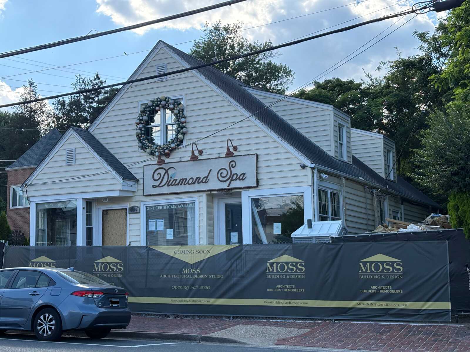 storefront with a white clapboard building reading 'Diamond Spa' above the entrance, framed by a festive circular wreath; construction fencing with 'MOSS Building & Design' banners in front and a parked gray car to the left