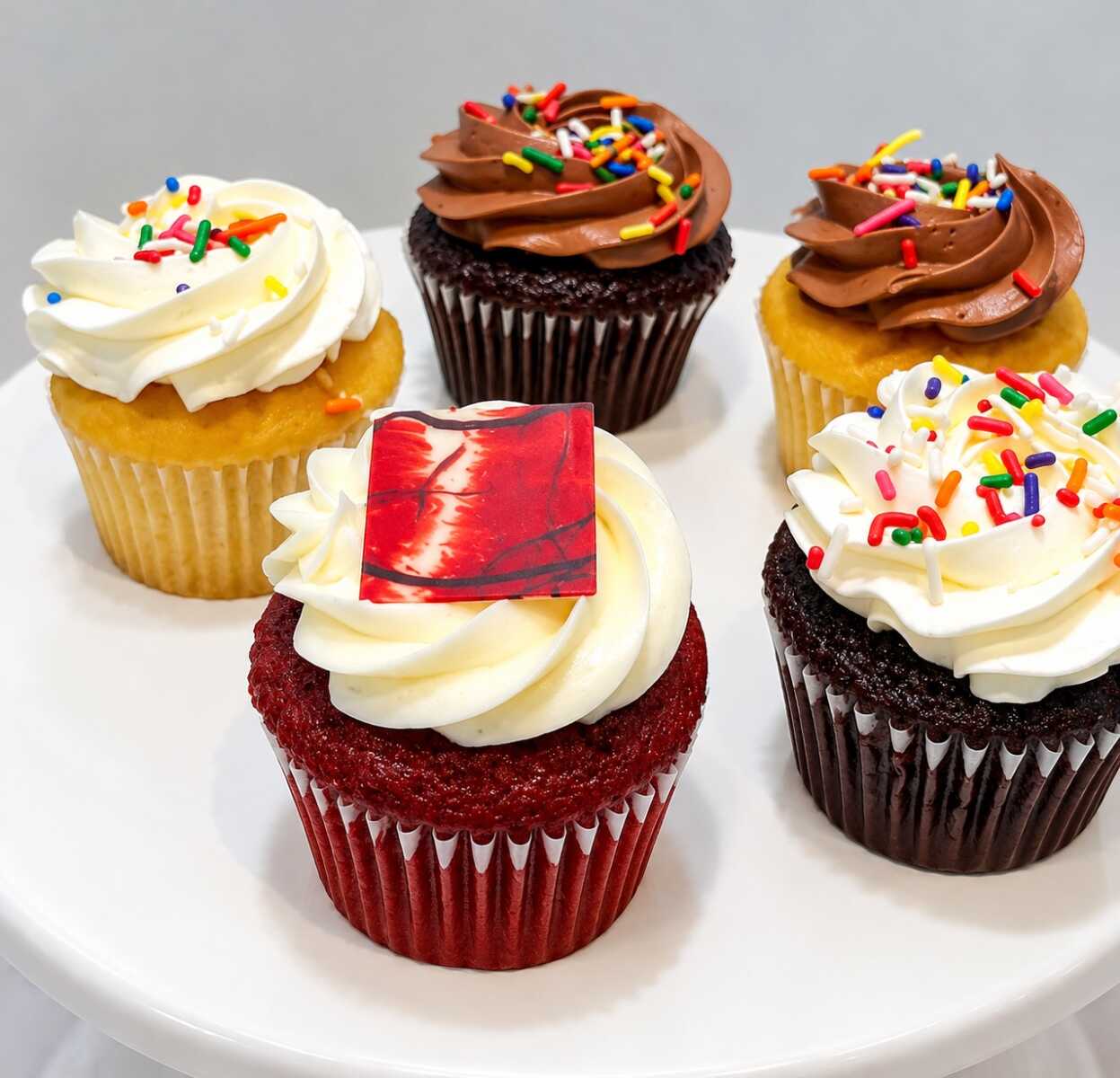 Assorted cupcakes with colorful sprinkles and swirled frosting on a white cake plate.