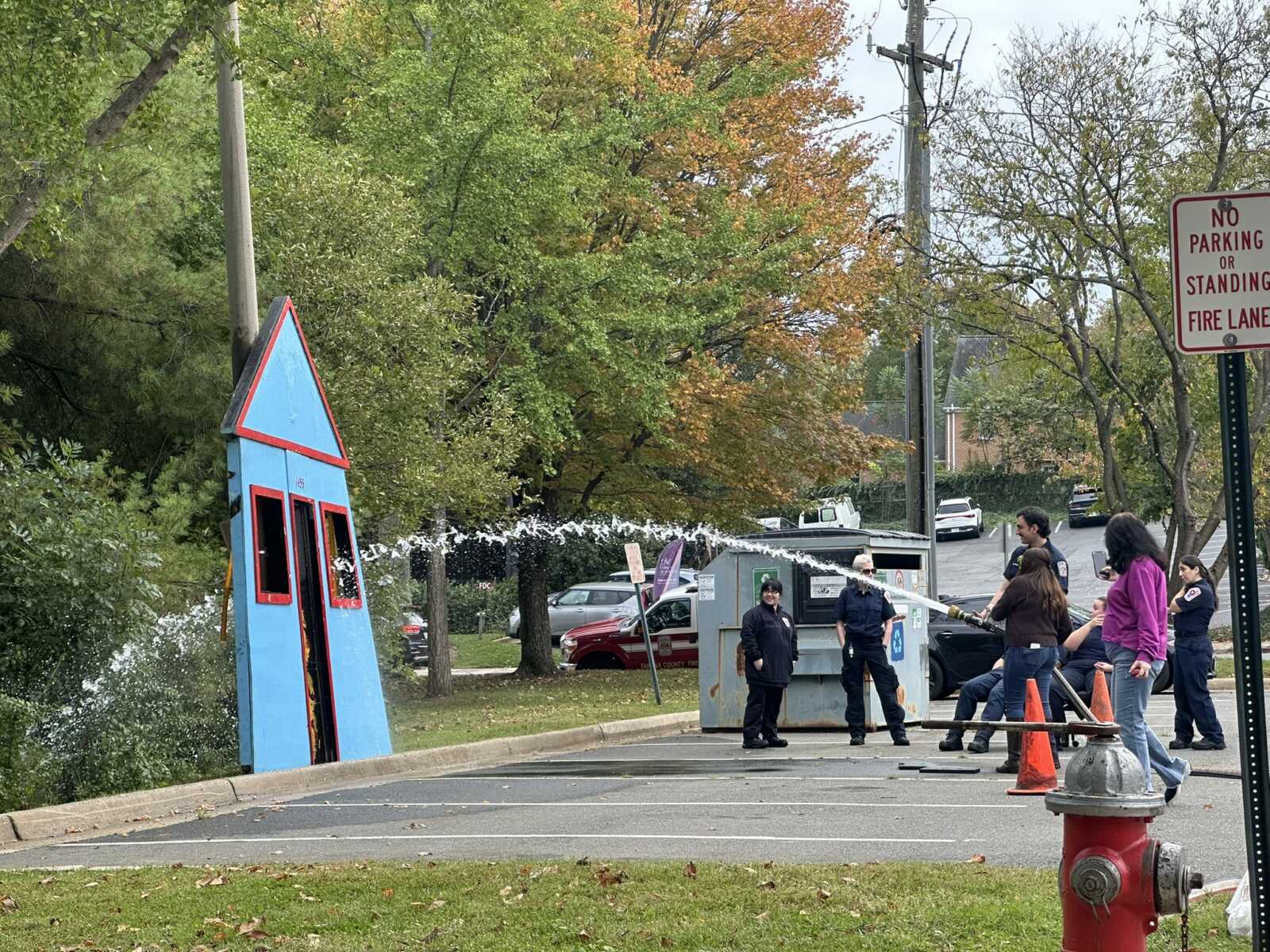 Emergency responders spray water from a hose at a toppled blue, house-shaped structure on a street while bystanders watch nearby.