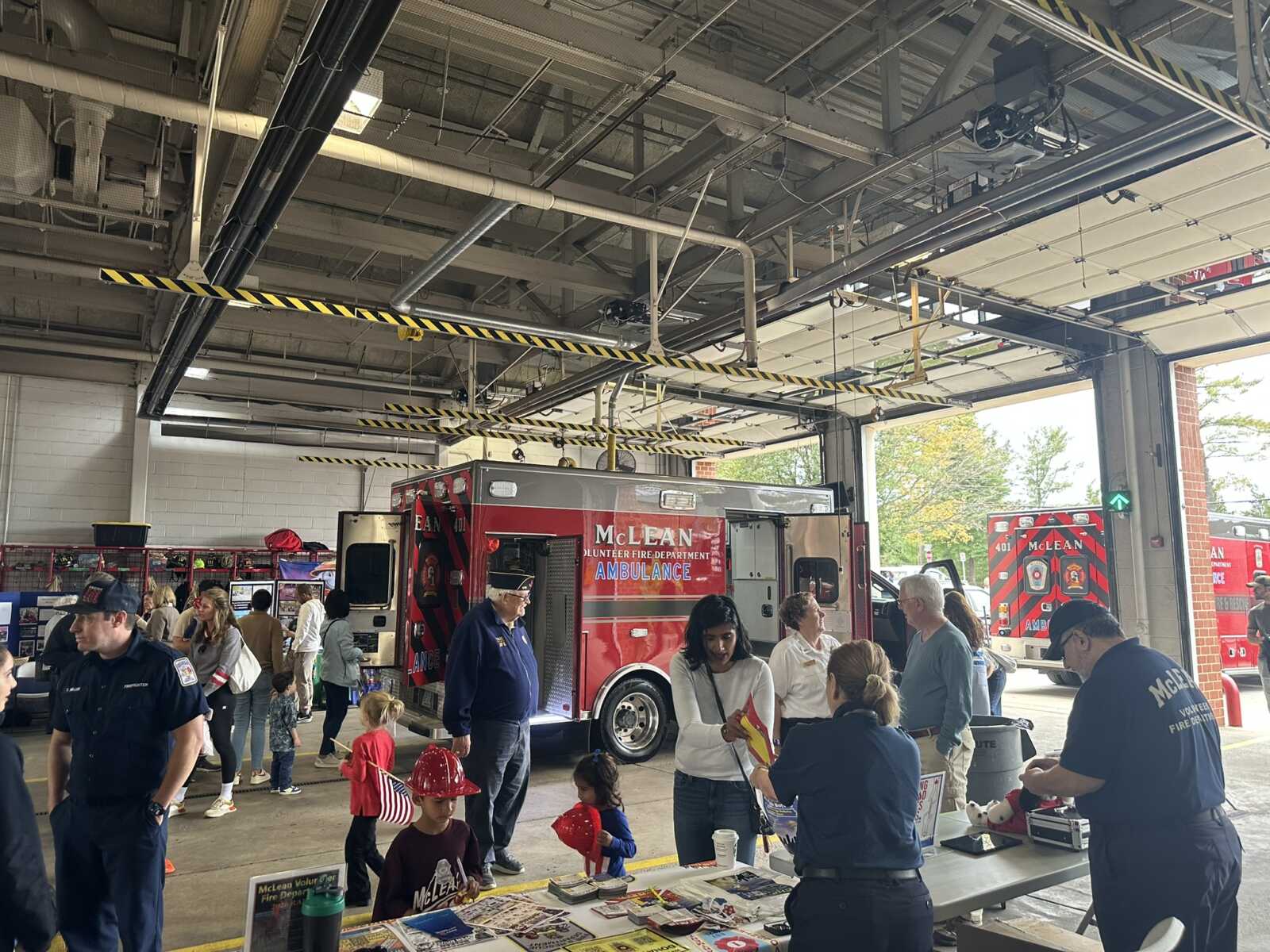 Indoor fire station open house with a red ambulance on display and families and kids exploring tables in the bay.