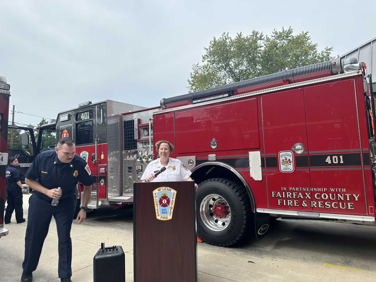 Female firefighter speaks at a podium in front of a large red fire engine during an outdoor briefing.