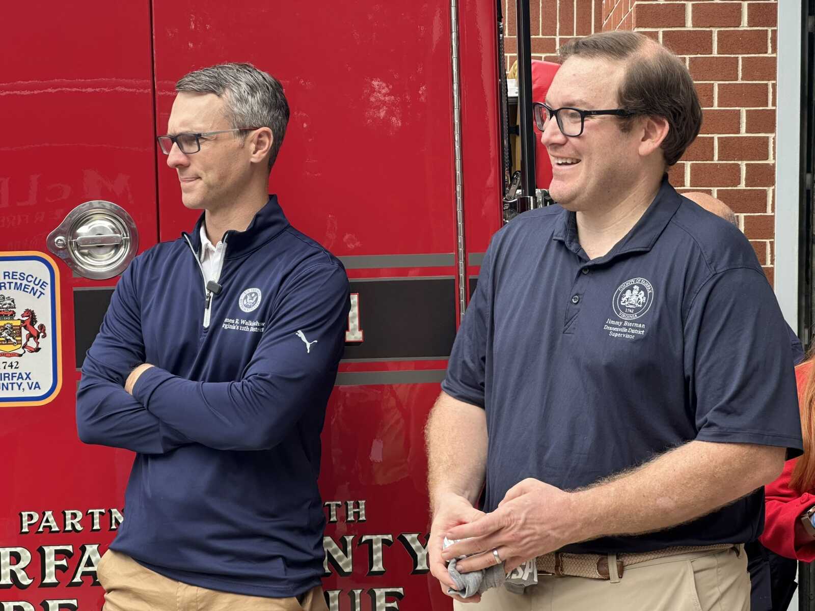 Two men in navy polo shirts stand beside a red fire truck, smiling and chatting outside a brick building.