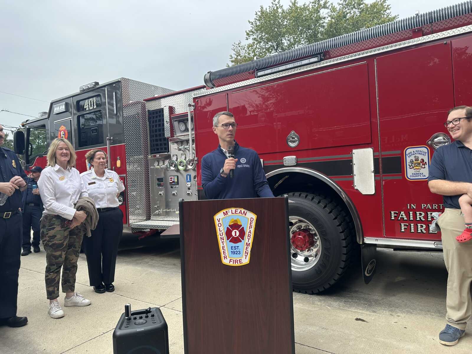 Speaker with microphone at a podium in front of a large red fire truck, flanked by uniformed staff at a community event.
