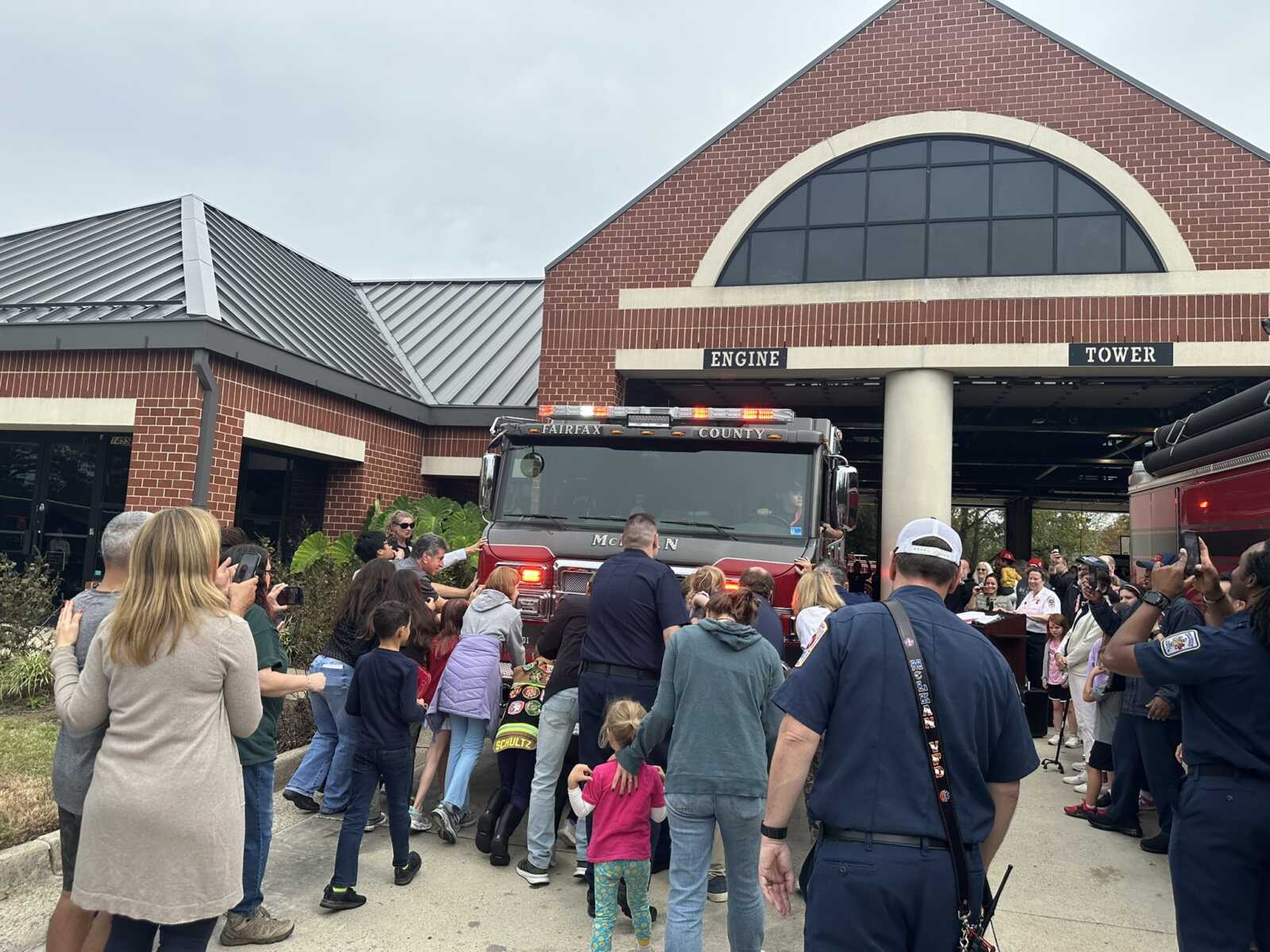 Crowd of families and kids gathered around a red fire truck parked outside a brick fire station labeled Engine and Tower.