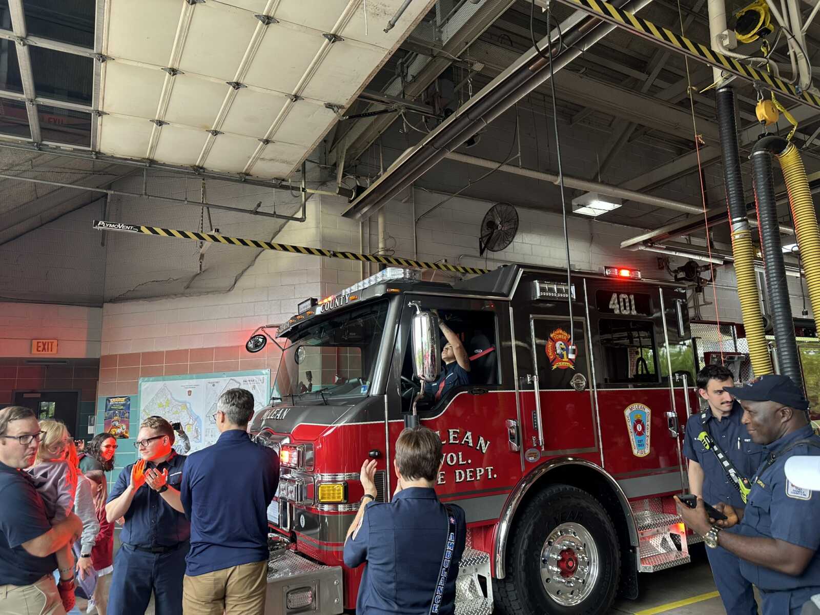 Red fire engine parked inside a station bay as firefighters and students chat nearby during a tour to the truck's side door open panels