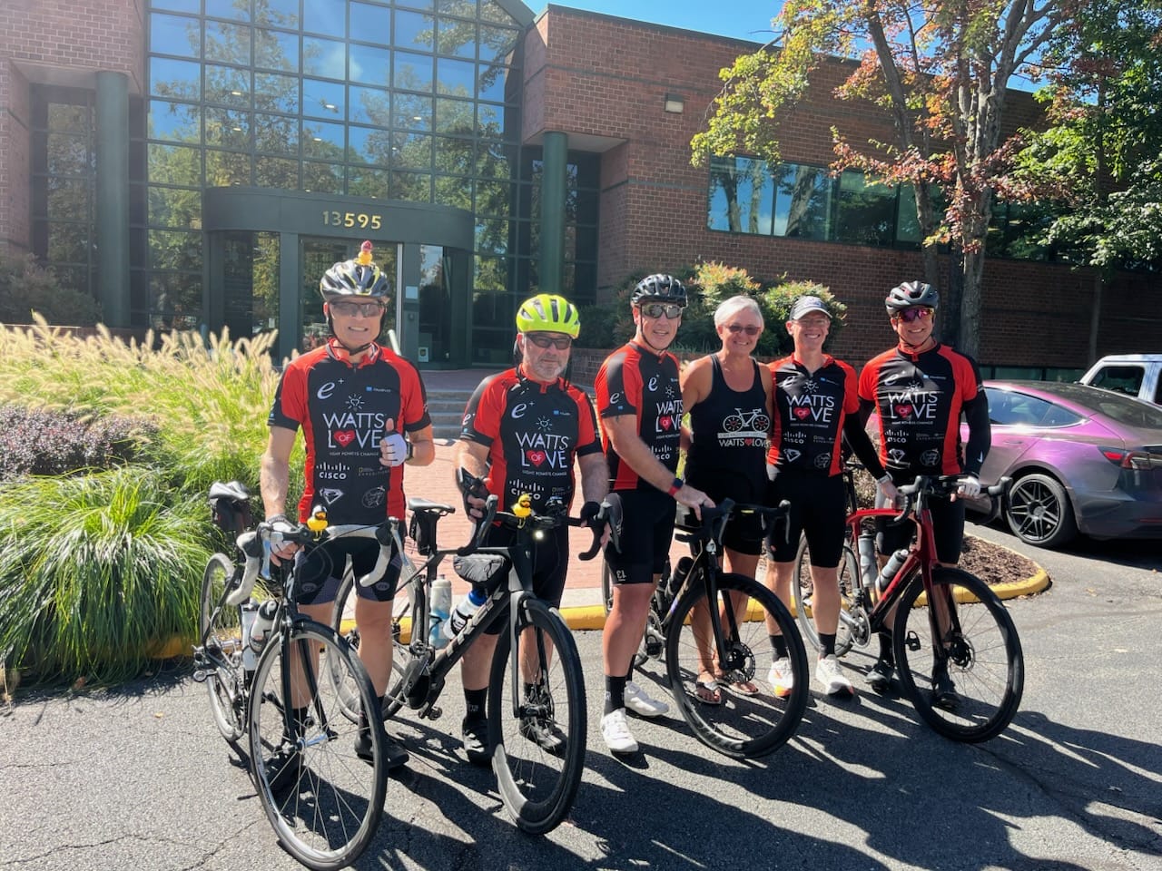 Group of six cyclists in red-and-black jerseys posing with their road bikes outside a brick office building with glass doors (address 13595).