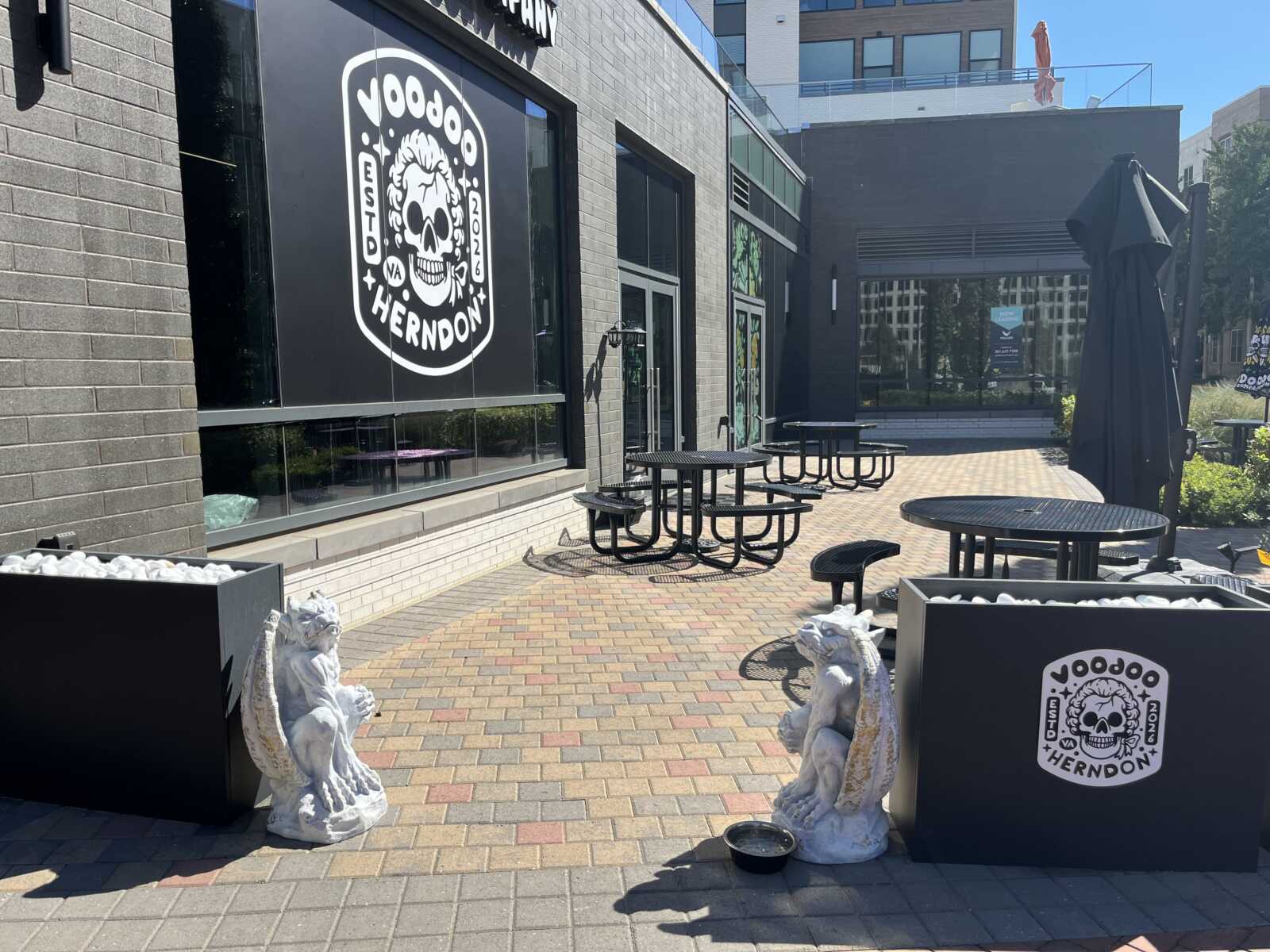 Outdoor dining area outside a restaurant/brewery with a bold skull logo reading 'VOODOO HERNDON' on a black wall, several round metal tables and benches, closed umbrellas, and decorative stone lions in planters under a sunny sky.