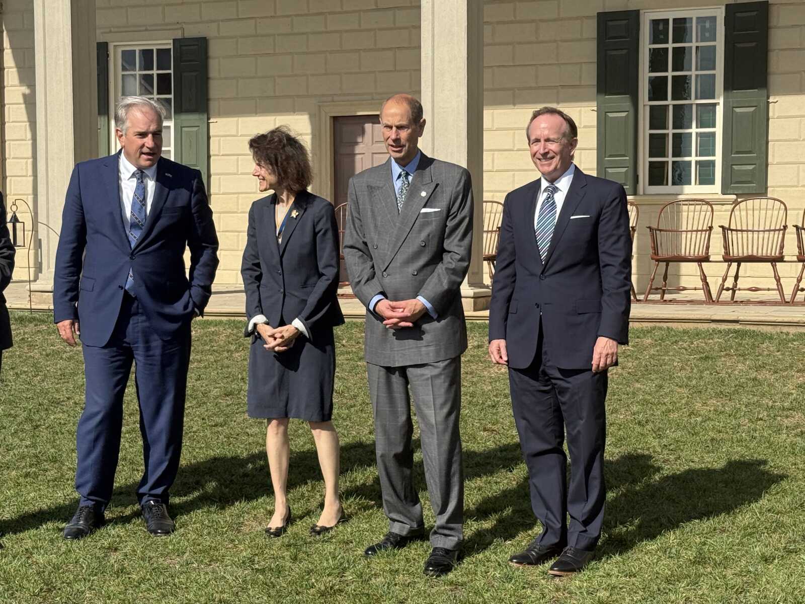 Four adults in business attire stand on a grassy lawn in front of a beige building with green shutters, posing for a photo.