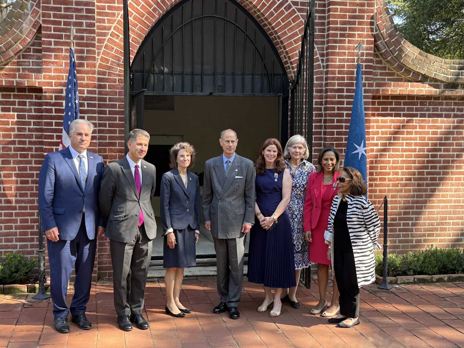 Eight professionally dressed individuals pose for a group photo in front of a brick archway with flag poles on either side.