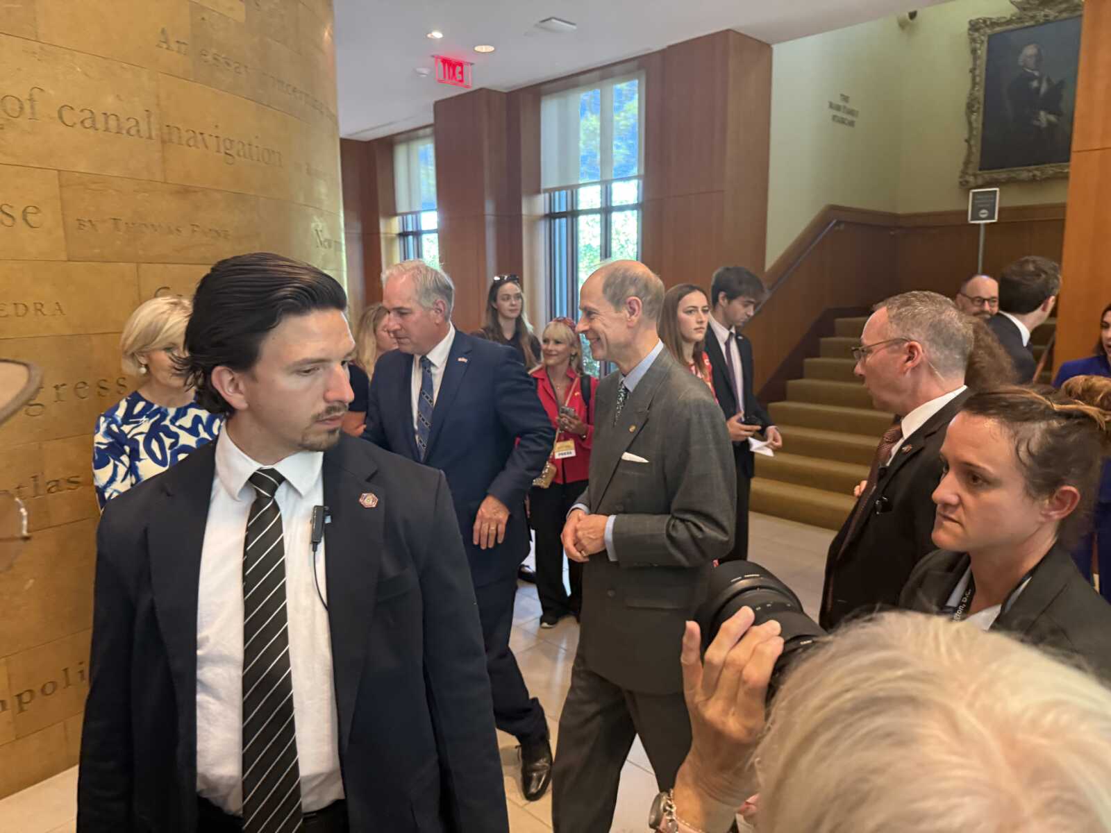 Man in a dark suit and striped tie walks past a stone wall in a formal lobby with people chatting in the background.
