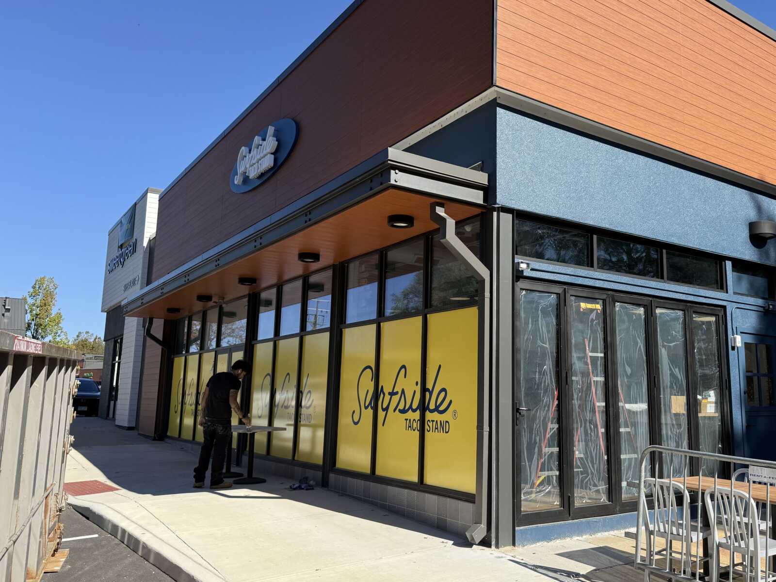 Exterior view of a modern storefront with orange and blue panels, large yellow window panels, and a person standing at a table outside.