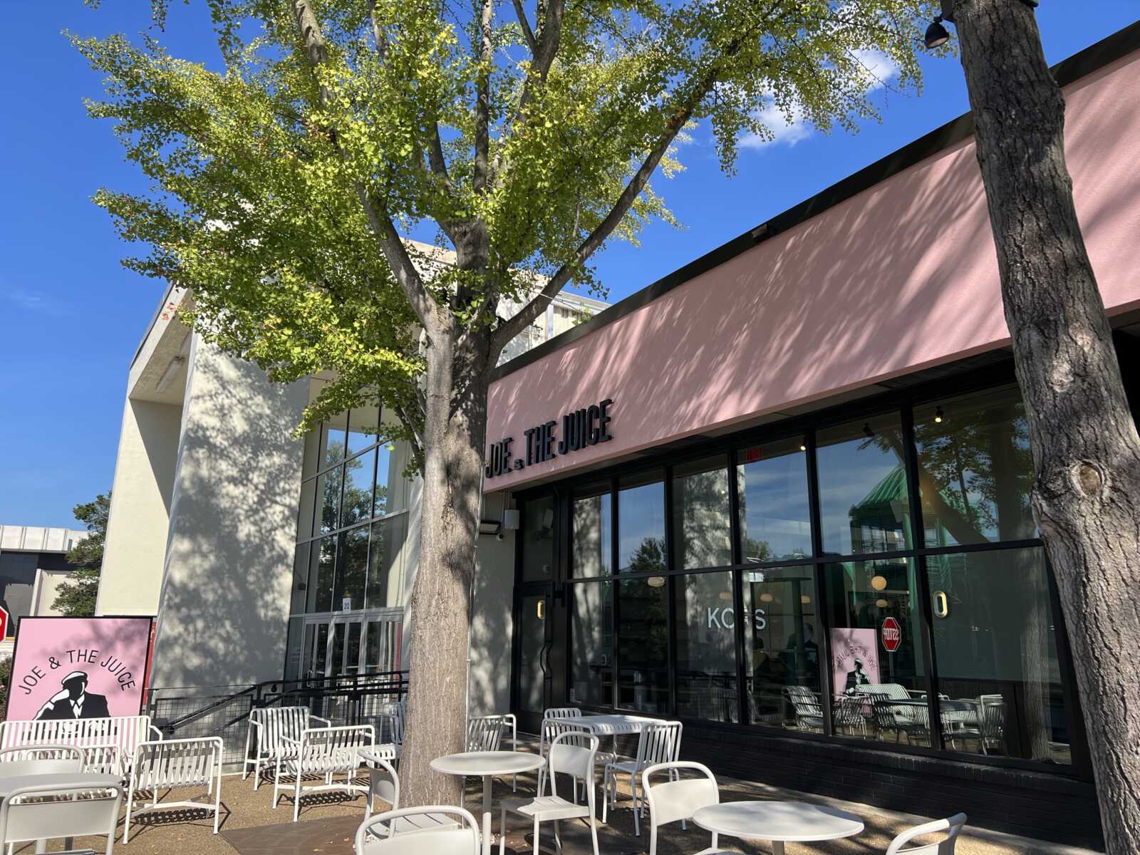 Outside Joe & The Juice store with a pink facade, large glass windows, and white outdoor tables beneath trees