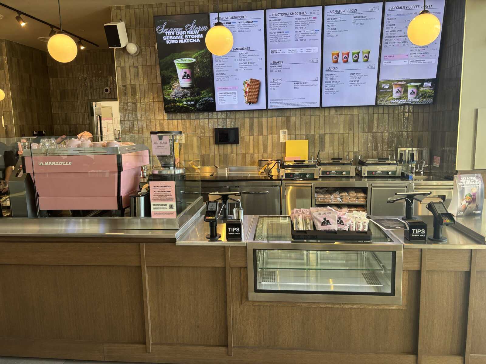 A cafe counter with a large pink espresso machine, glass display case, and digital menu boards above the back wall.