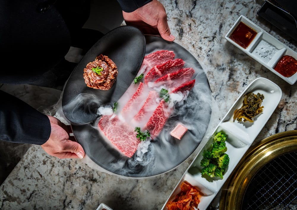Chef presenting thinly sliced marbled beef on a black plate with a smoky effect, surrounded by dipping sauces and vegetables on a marble table.