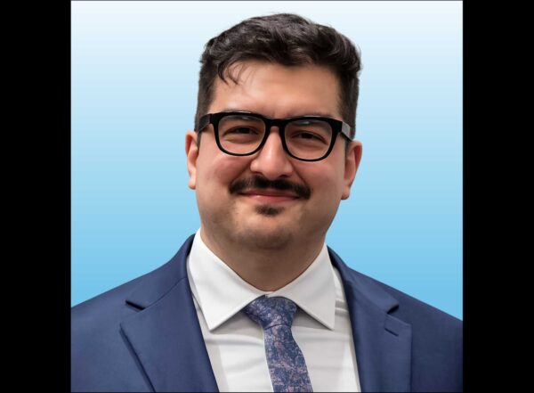 Professional head-and-shoulders portrait of a man with dark hair and glasses, wearing a navy suit and patterned tie against a light blue gradient background.
