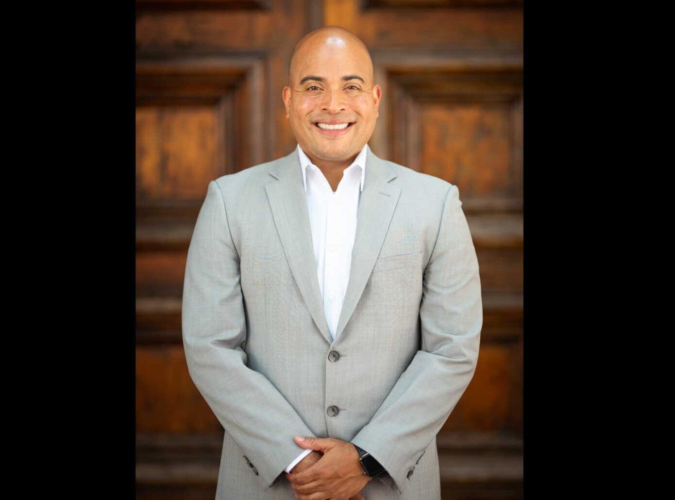 Headshot of Herndon Town Council member Kelvin Garcia (photo by PJ Morgan) | FFXnow Smiling man in a light gray suit and white shirt stands with hands clasped, against a dark wooden paneled backdrop.