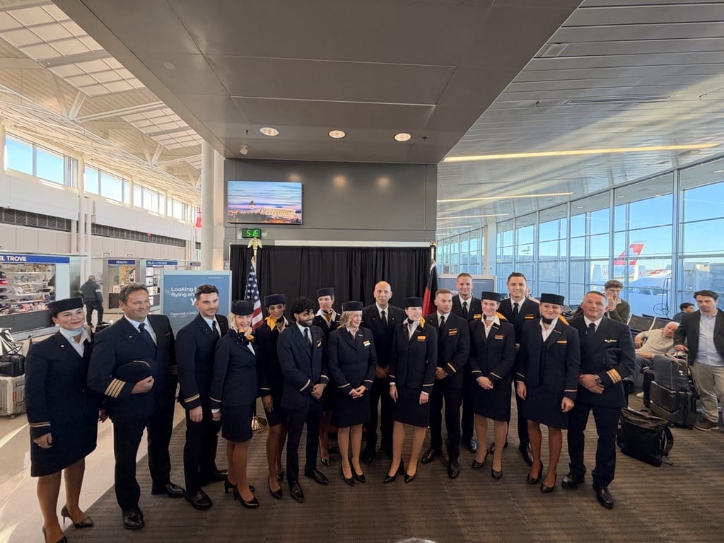 Group of airline crew in uniform posing for a photo at an airport terminal with an American flag in the background.