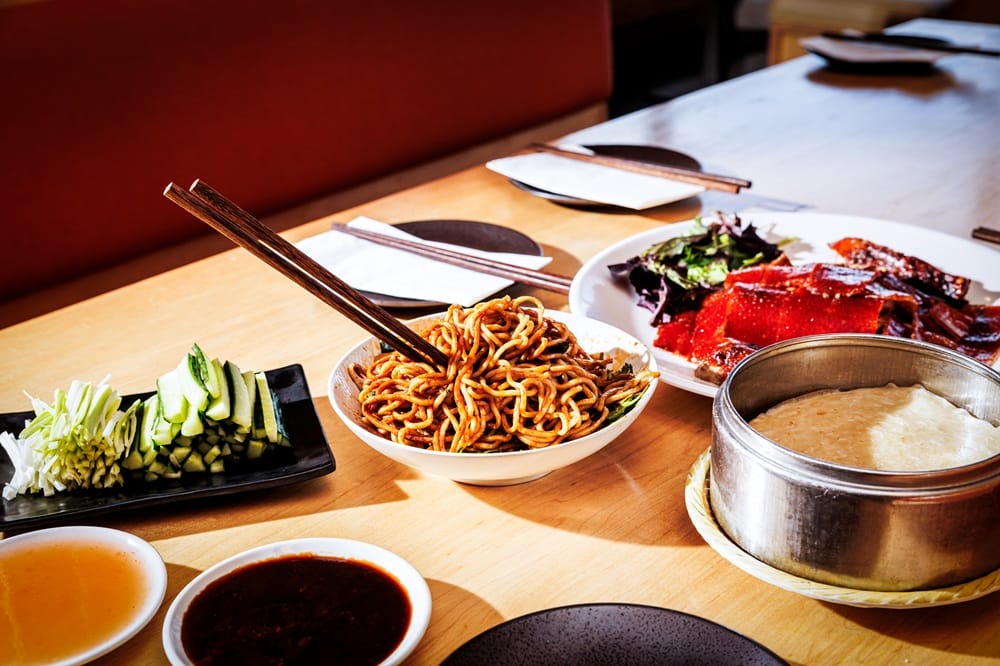 Assorted Asian dishes on a wooden table: noodles, cucumber sticks, greens, sliced meat, and a pot of rice with sauces at a restaurant table