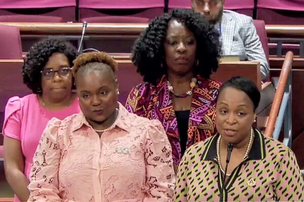 Four women stand at a lectern in a formal setting; one speaks into a microphone behind them right now.