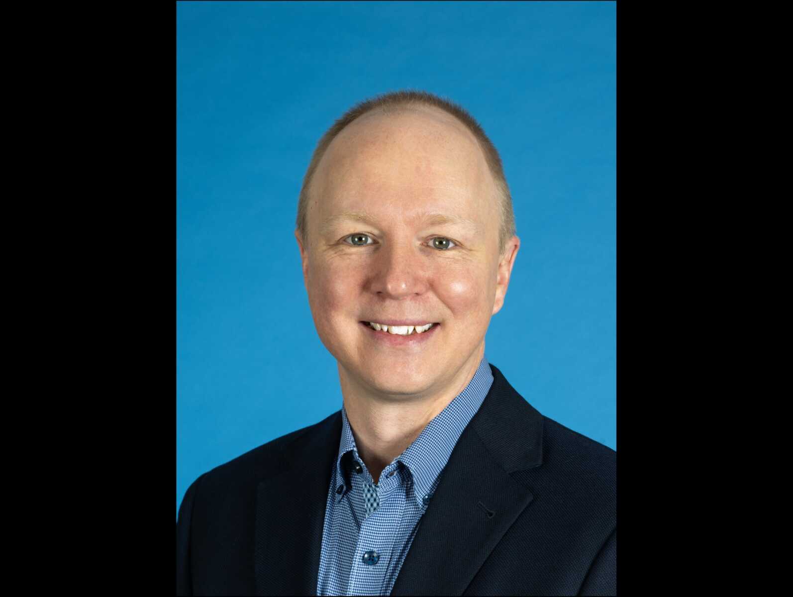 Matthew Mullenix (courtesy of Reston Association) | FFXnow Smiling man in a dark blazer and blue checkered shirt, head-and-shoulders portrait against a blue backdrop