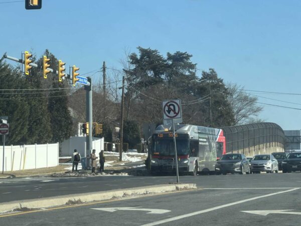Bus at a busy intersection with cars lined up and pedestrians nearby on a blue-sky day. A no U-turn sign is visible near the curb.