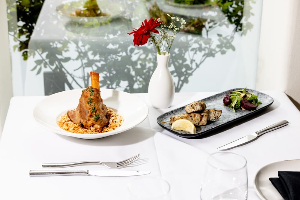 Fine dining table with two dishes: lamb shank on risotto and grilled fish with greens, white vase with red flower in background.