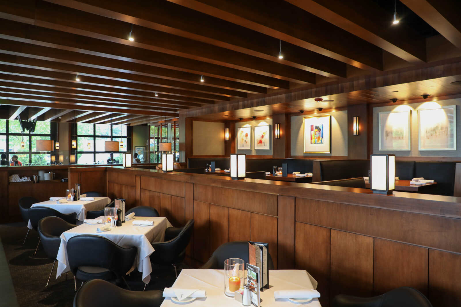 Elegant restaurant dining area with white-clothed tables, dark chairs, and warm candlelight under wooden beam ceiling.