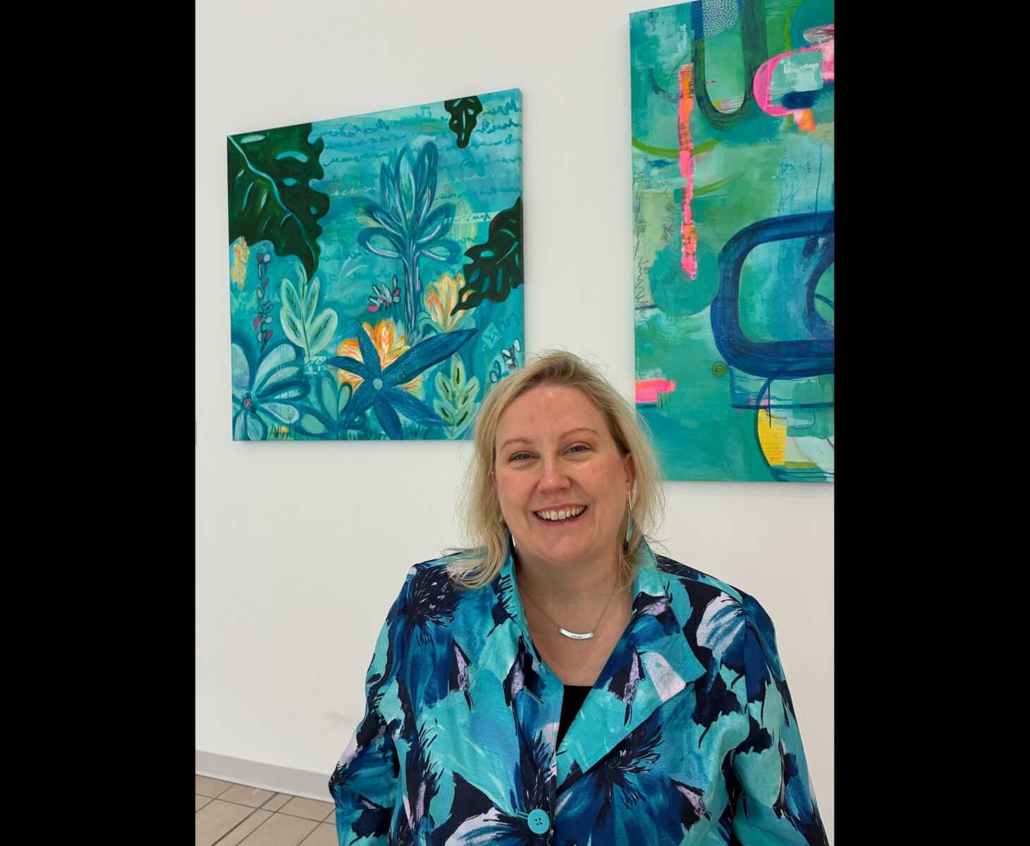 Precious Burger with paintings from her new exhibit "Through Me: Where Wonder Finds a Way" at Springfield Town Center (courtesy Springfield Town Center) | FFXnow Smiling woman with blonde hair in a turquoise floral jacket, standing in front of blue abstract paintings.