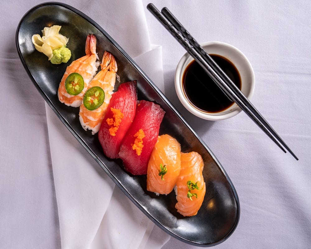 Assorted nigiri sushi on a black oval plate with salmon, tuna, and shrimp, plus wasabi and pickled ginger, with soy sauce and chopsticks nearby.
