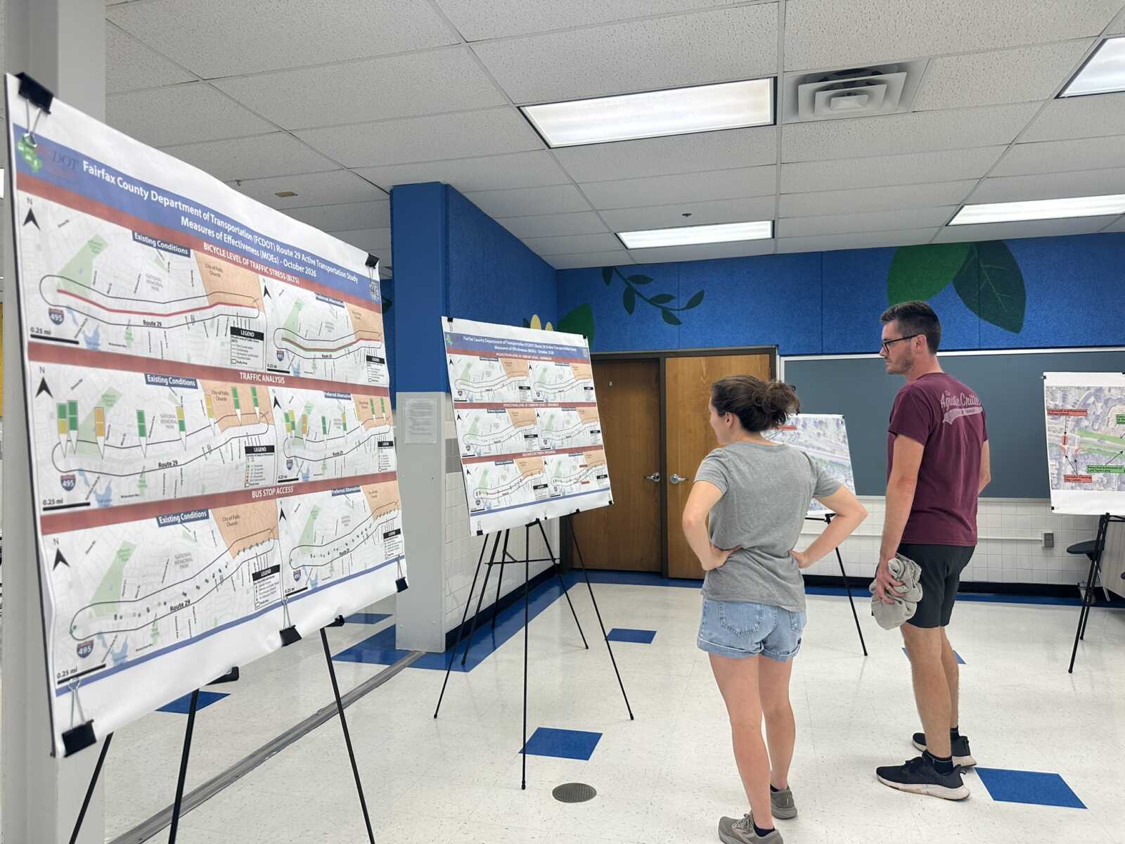 Two people study large traffic maps displayed on easels in a bright room with blue walls and a tiled ceiling.