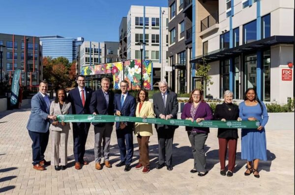 A group of 10 men and women stand behind a ribbon, with housing units visible behind them.