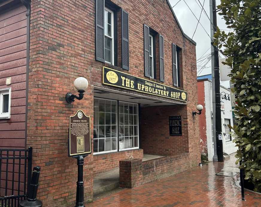 Brick storefront of The Upholstery Shop with a black-and-gold sign, large front window, and brick sidewalk outside.
